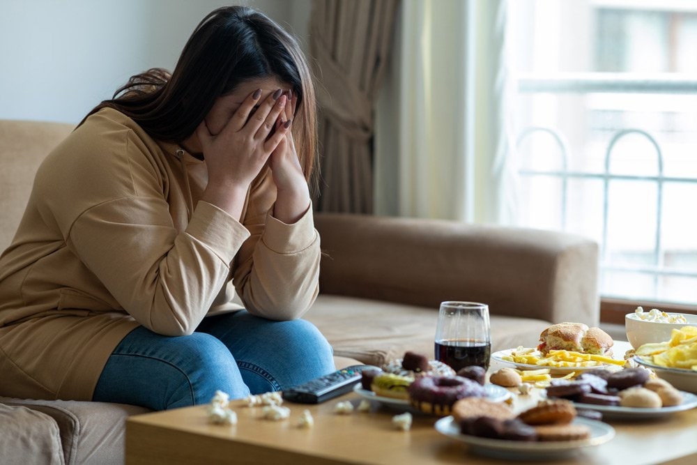 photo of a distress woman sitting on a couch in a living room in front of a table full of food