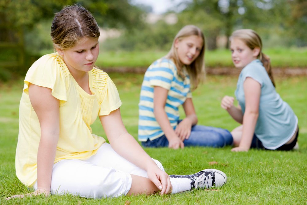 photo of a girl sitting on grass while 2 other girls judging her from a distance